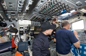 Two astronauts inside the flight deck monitor through the windows and communicate to the astronauts conducting a spacewalk.