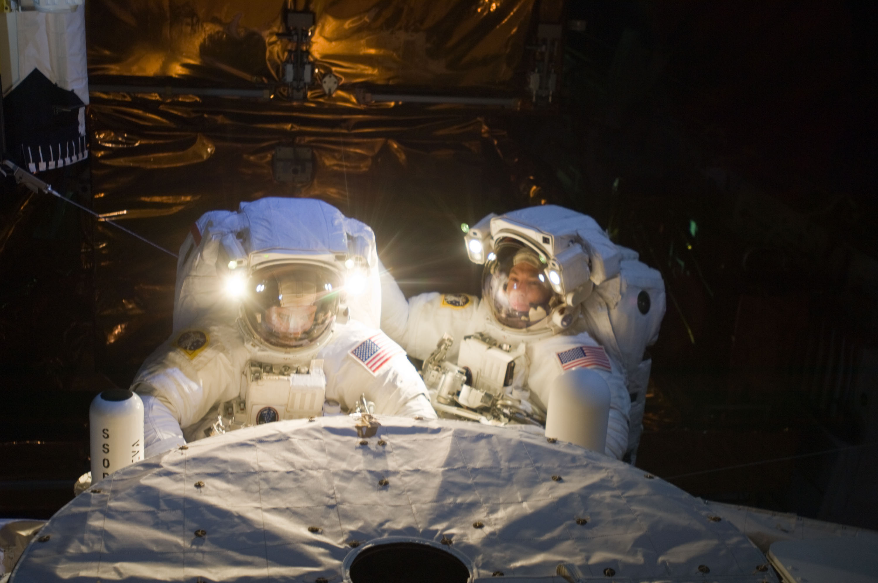Two astronauts at the airlock on the shuttle with their helmet lights turned on.