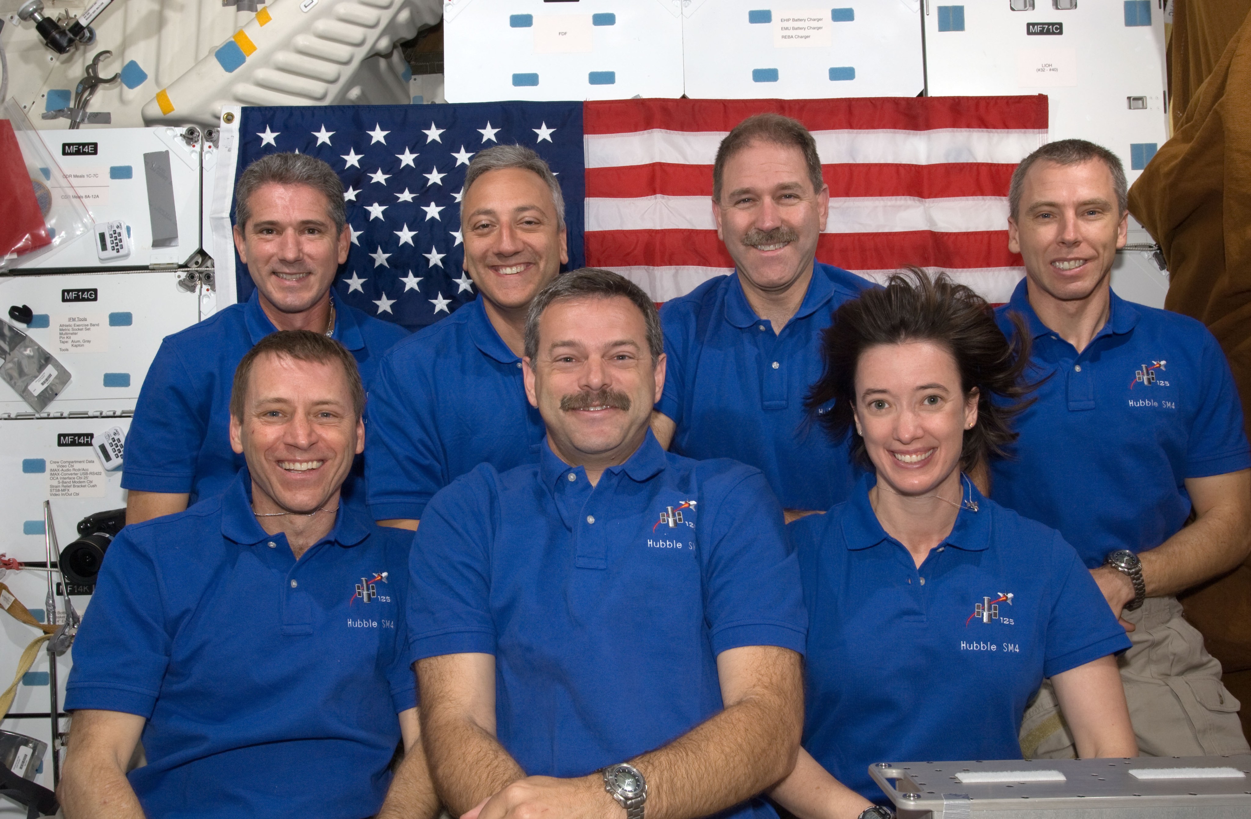 STS-125 crew poses in front of an American flag in the crew cabin of the shuttle.