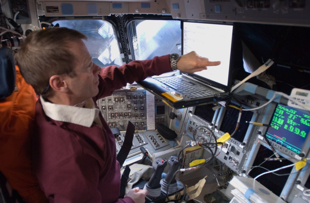 The shuttle pilot at the pilot's station points at something on a laptop screen.