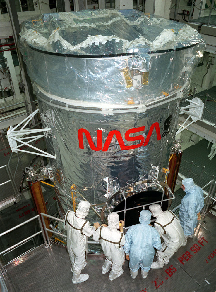 Engineers in cleanroom suits gather around the top of Hubble.