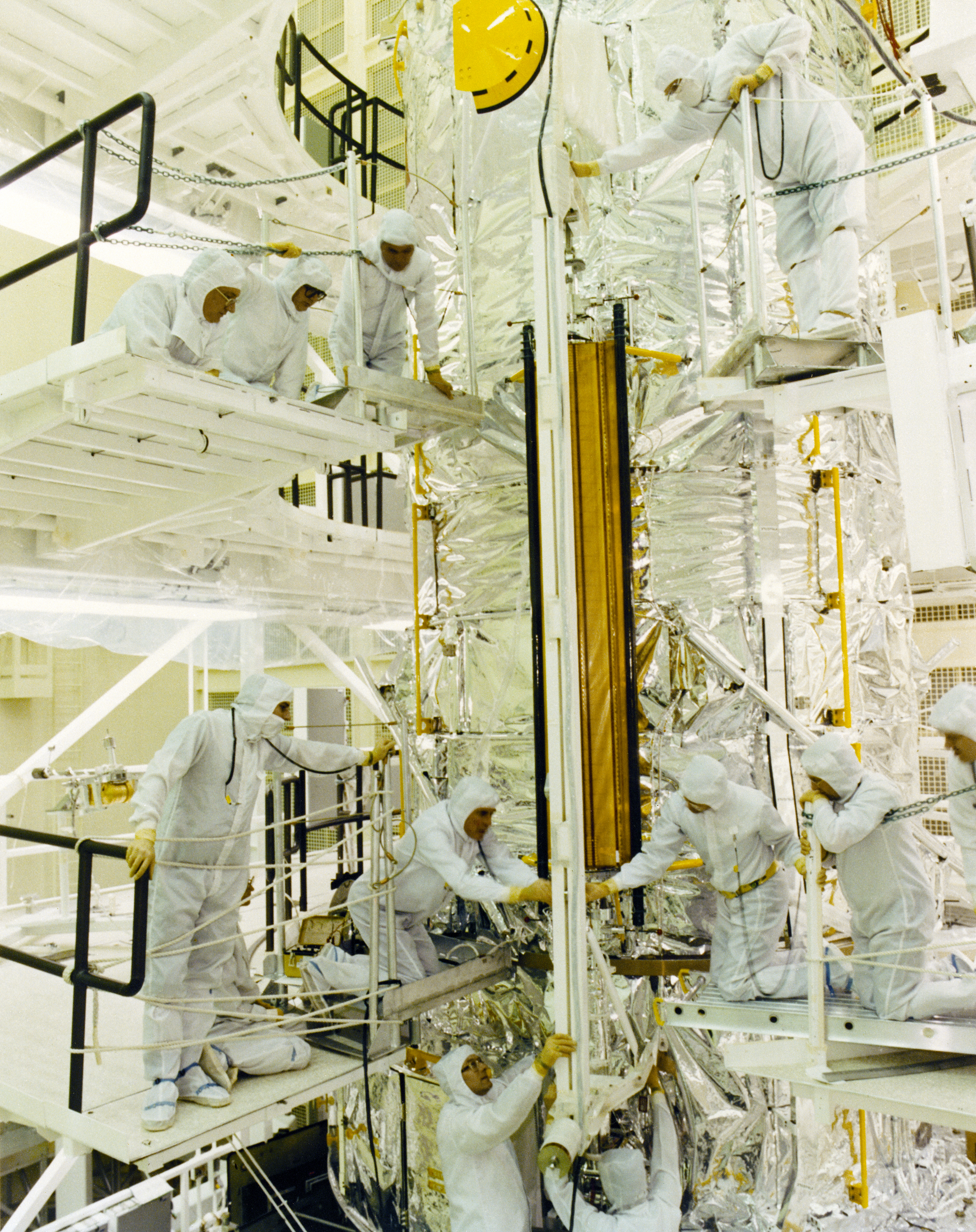 Engineers in cleanroom suits gather around the solar array attached to Hubble at multiple heights.