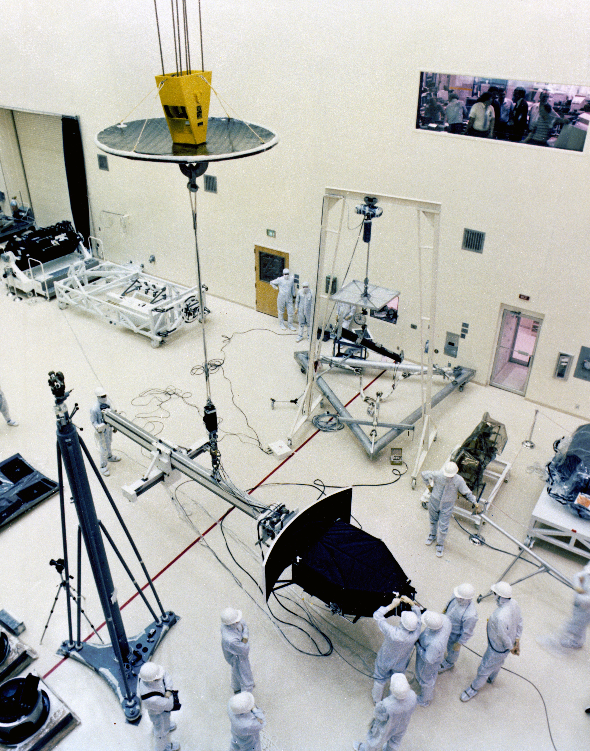The Wide Field Planetray Camera is being lowered on a crane for installation in a clean room surrounded by engineers.