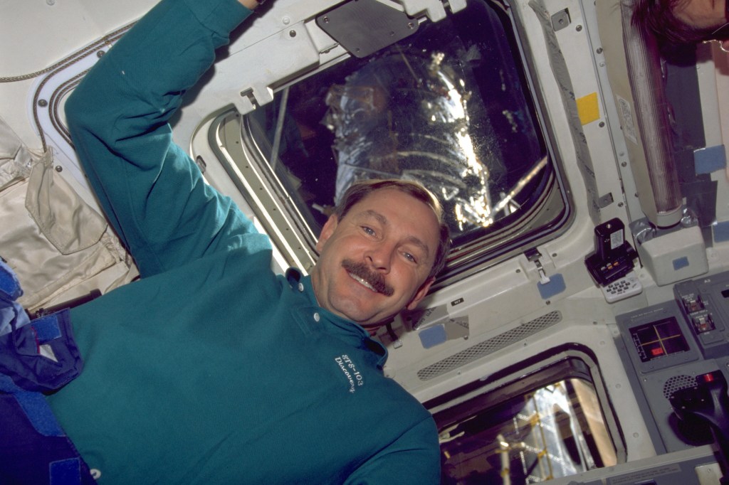 An astronaut floats in front of the windows of the shuttle from inside the crew cabin. Hubble can be seen through the window.