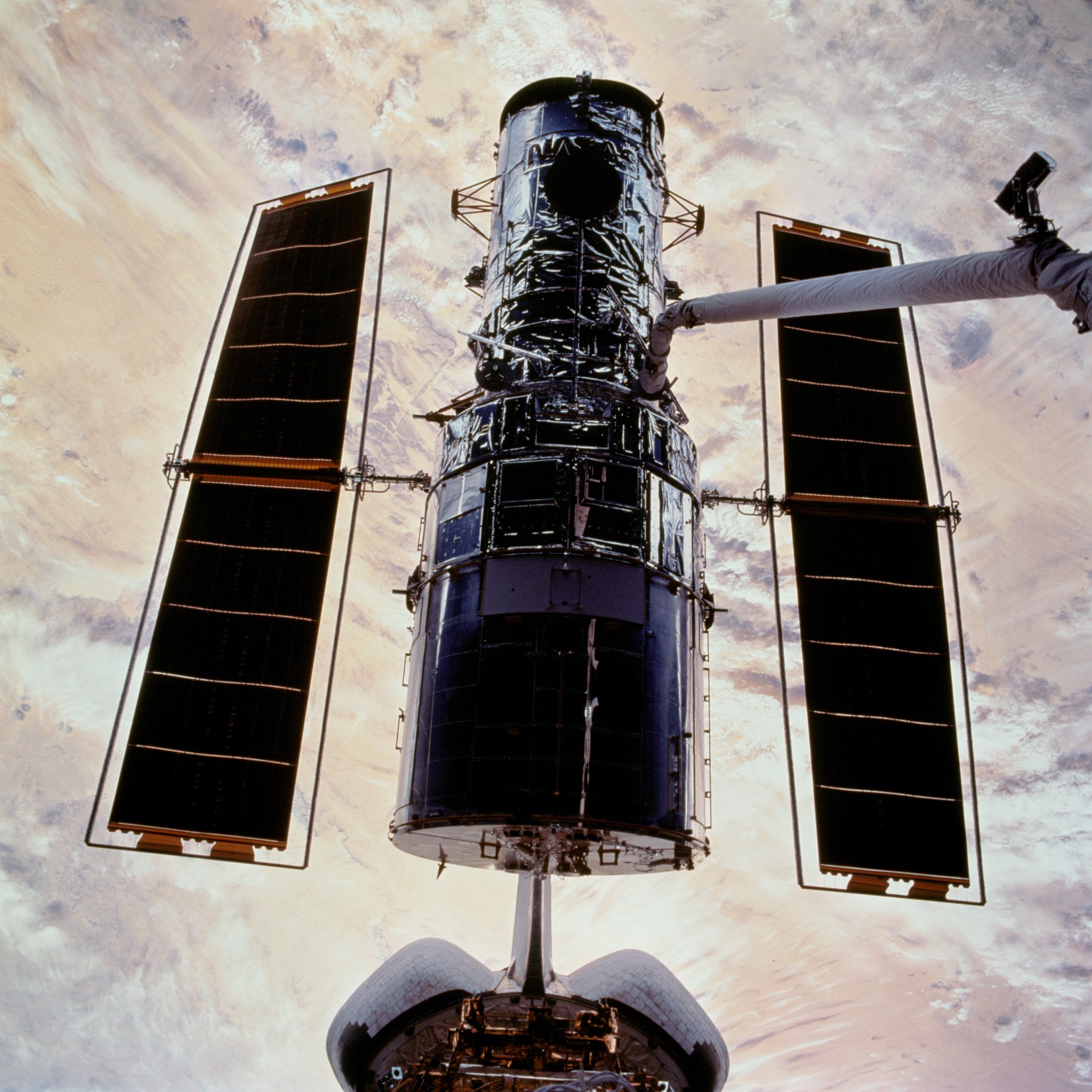 Hubble, on the end of the robotic arm after it was captured in space, is lowered onto its berthing hardware in the cargo bay, with a colorful section of earth below.