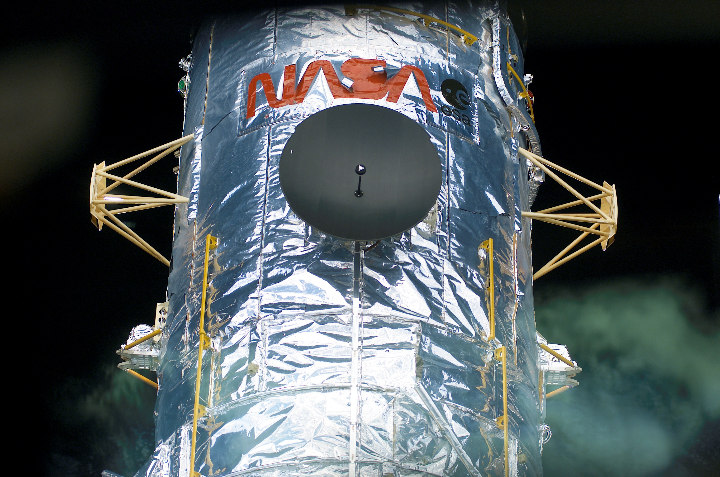 The top portion of the Hubble Space Telescope is seen in this image, with the red NASA worm logo on it, against black space.