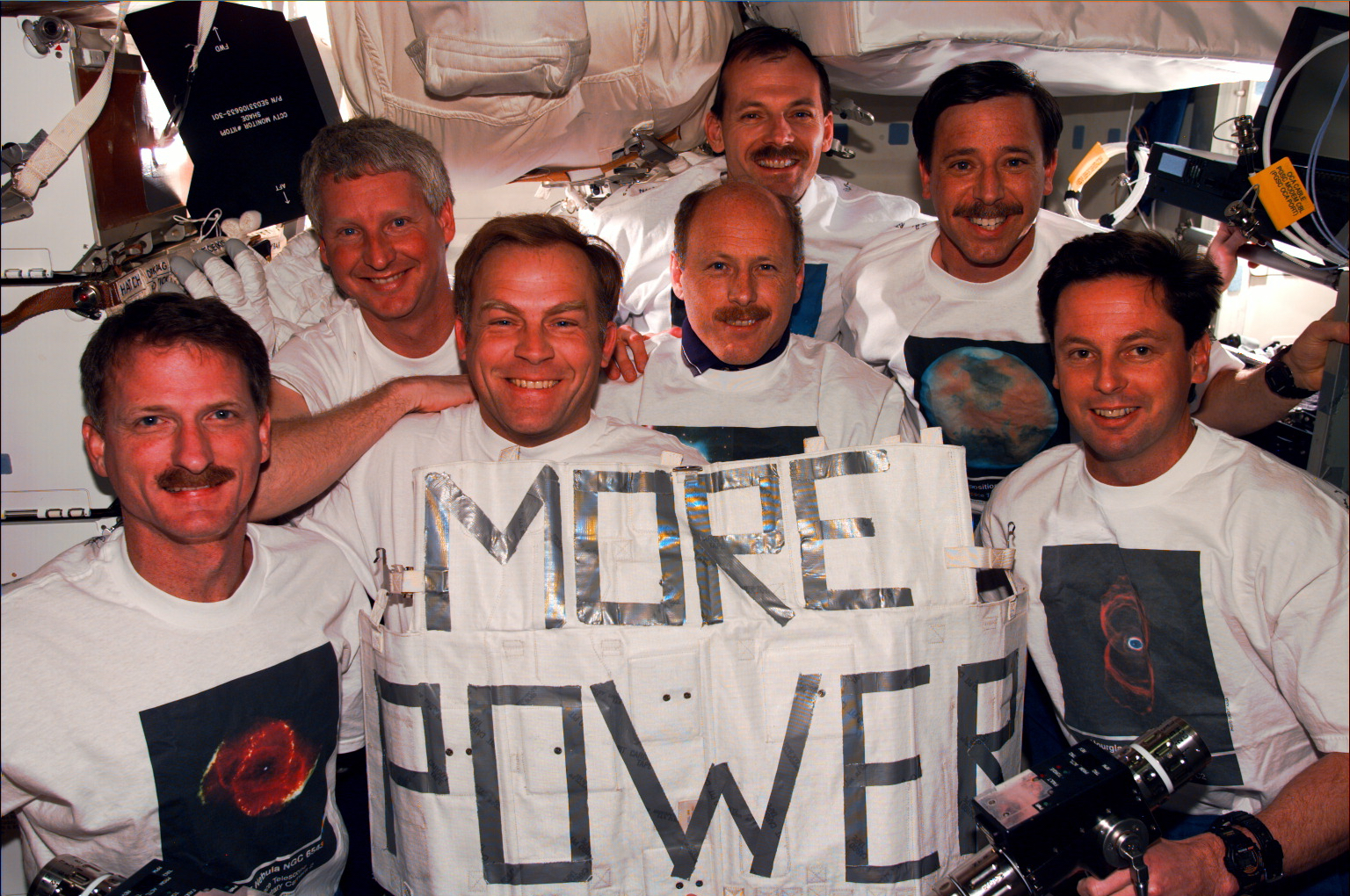 A crew photo inside the shuttle's cabin with them holding a sign that says "MORE POWER"