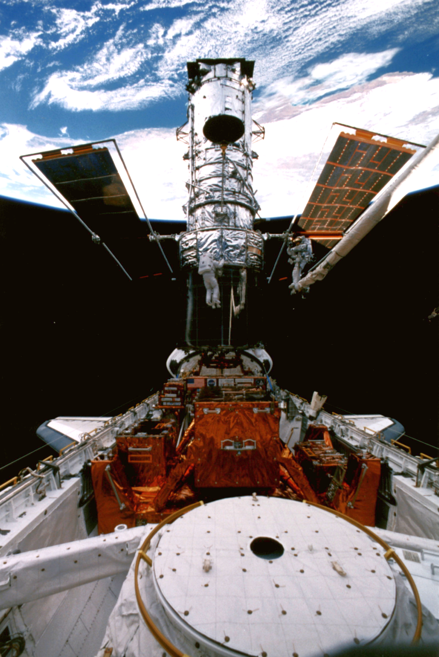The Hubble Space Telescope docked in the cargo bay of the shuttle with astronauts working on it. A portion of the blue earth can be seen behind it.