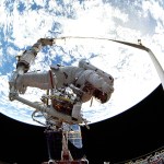 An astronaut on th end of the robotic arm with many tools attached to a workstation and a large blue earth in the background.