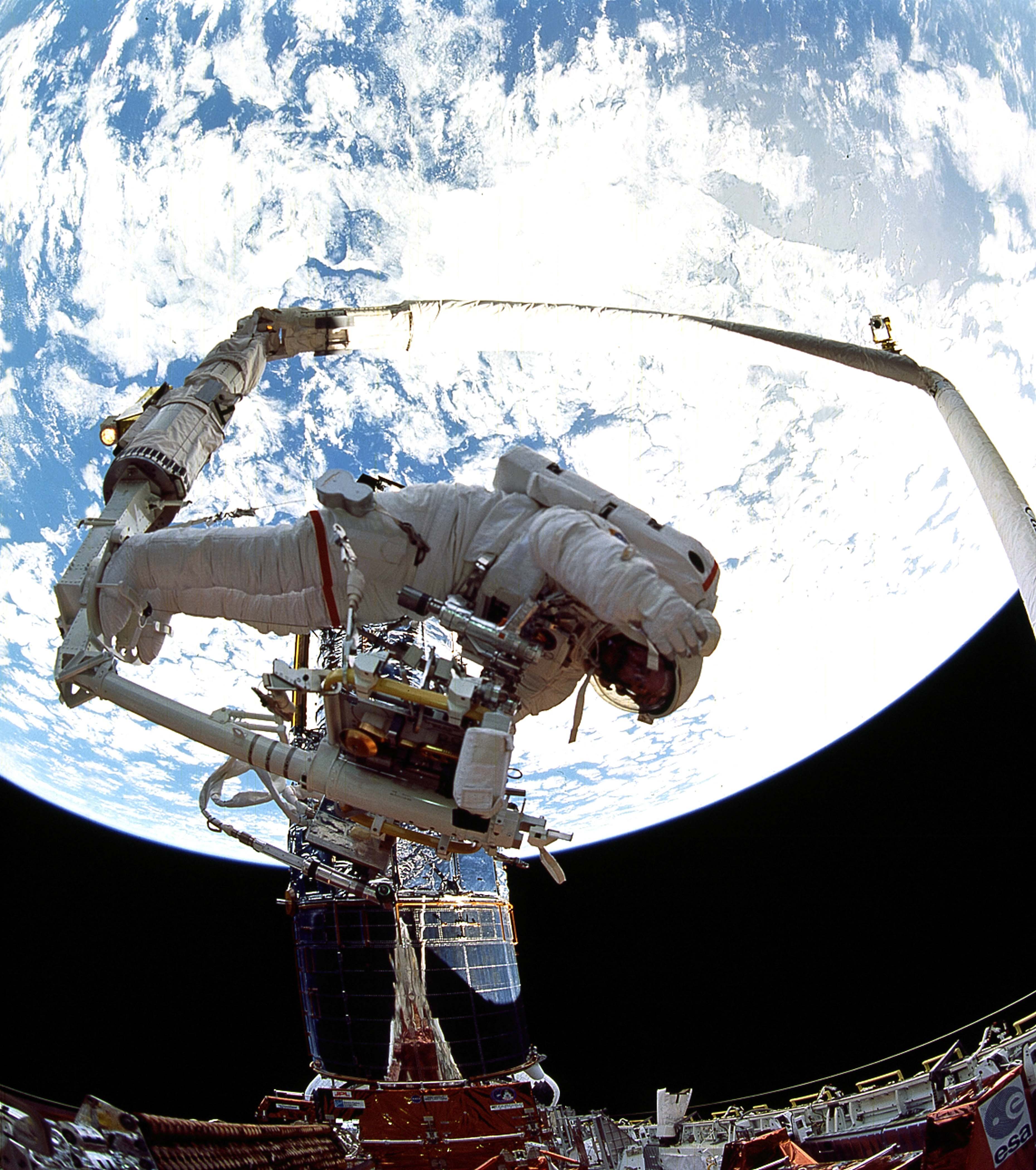 An astronaut on th end of the robotic arm with many tools attached to a workstation and a large blue earth in the background.