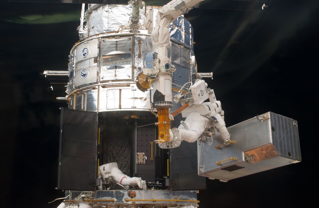 Two astronauts working on Hubble. One has an instrument (COSTAR) in his hands while the other is inside the bottom of the spacecraft.