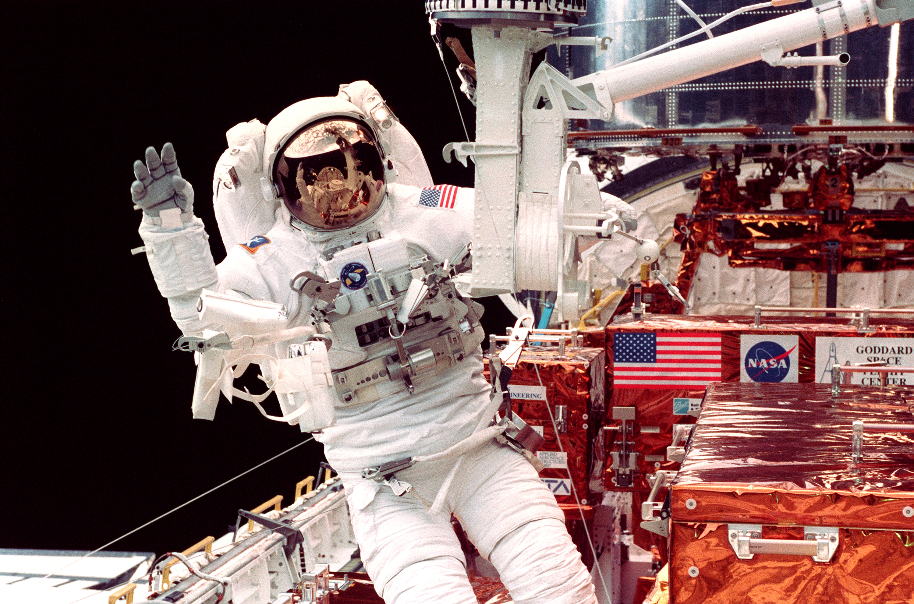 An astronaut waves to the camera in the cargo bay of the space shuttle.