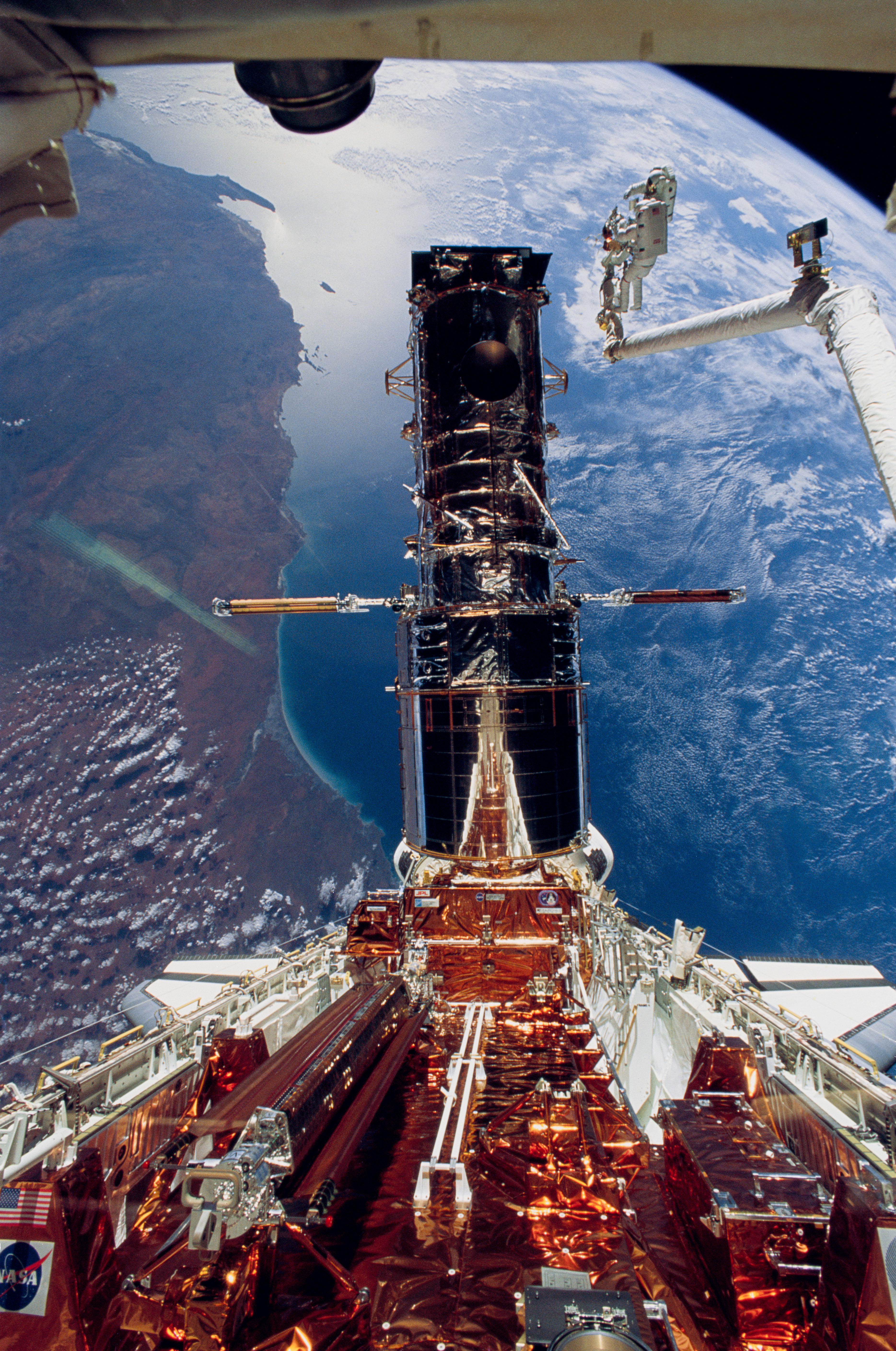 Two astronauts on the end of the shuttle's robotic arm are raised to the top of Hubble. A full Hubble, and the gold cargo bay carriers are seen in front of earth.