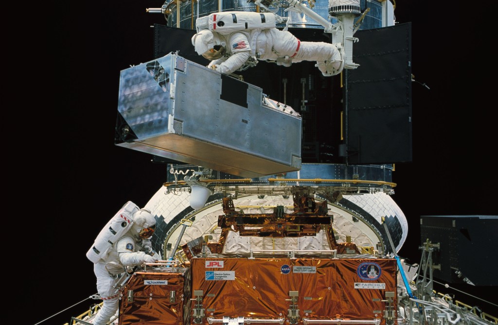 An astronaut on the robotic arm of the shuttle carries the COSTAR instrument for installation whil another works in the cargo bay.