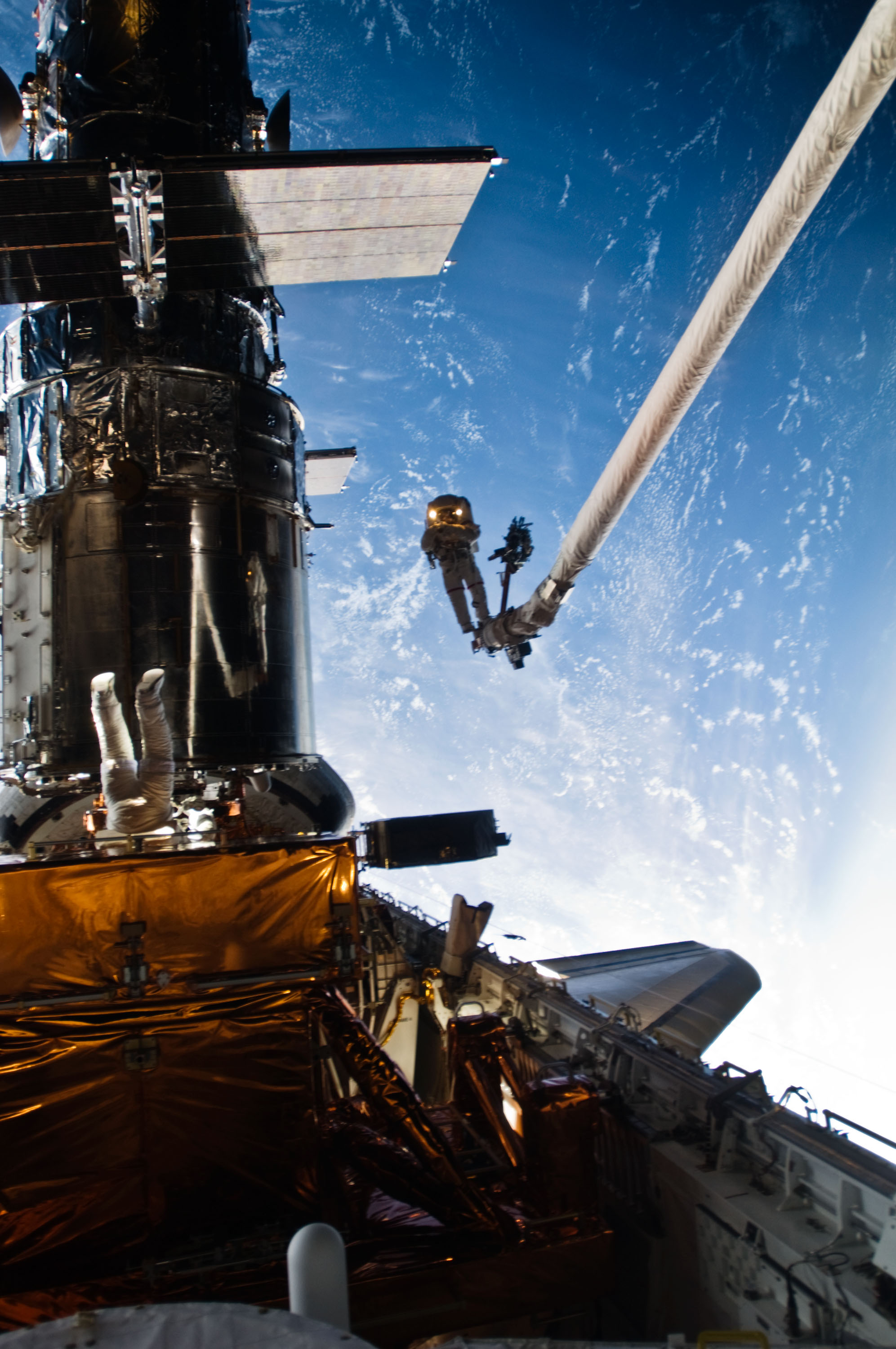 An astronaut silhouetted against the earth with a second astronaut upside down near a cargo bay carrier.