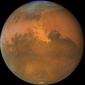 Full shot of Mars. Rusty red, you can see many of the ground features of the planet, with dust coming from the north.