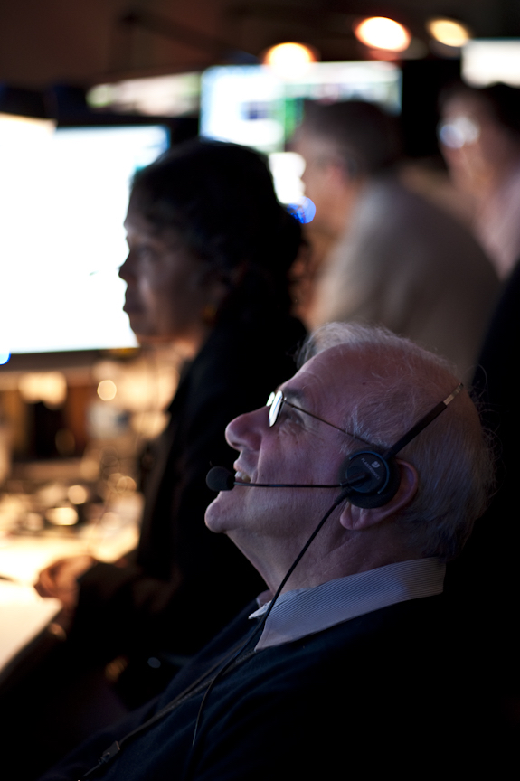 A person with a headphone and microphone on his head watches the mission on a screen in front of him.