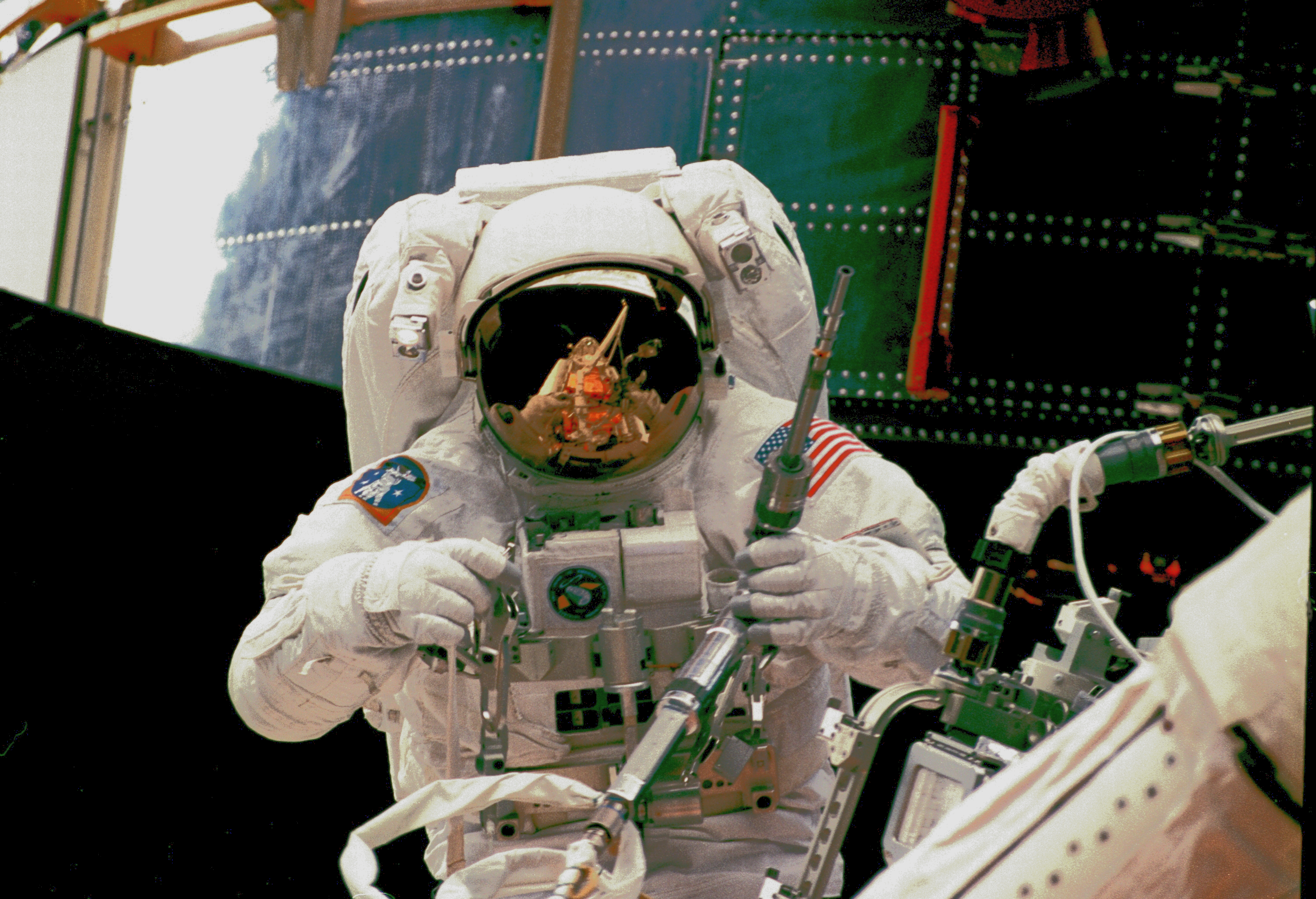An astronaut with a power ratchet in one hand in front of Hubble. The shuttle cargo bay, gold carriers, and robotic arm are reflected in his visor.