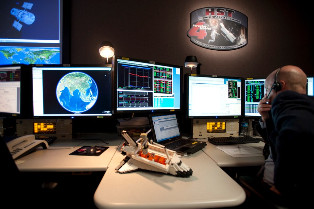 A LEGO model of the Shuttle, cargo bay carriers, and Hubble on a robotic arm sits next to the montiors in the Hubble control center.