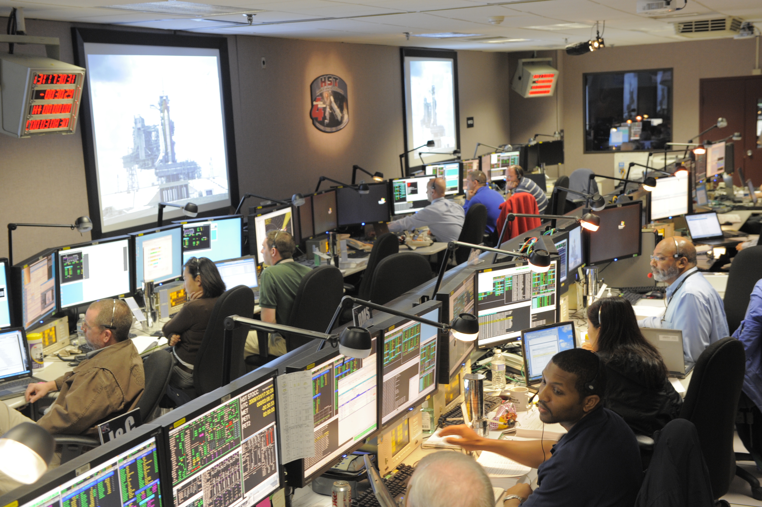 Hubble's Operations Support Room in the control center is fully staffed at consoles while the shuttle on the launch pad is on the big screens.
