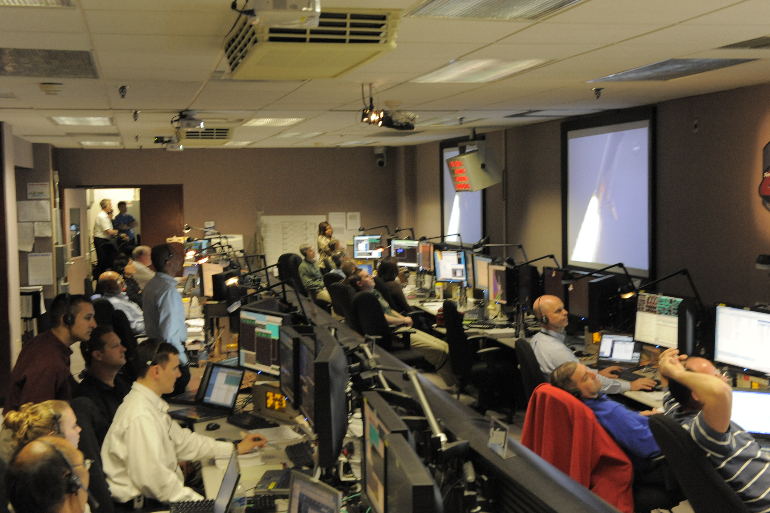 A fully staffed Hubble control center room watches the launch of a shuttle on the large screens.