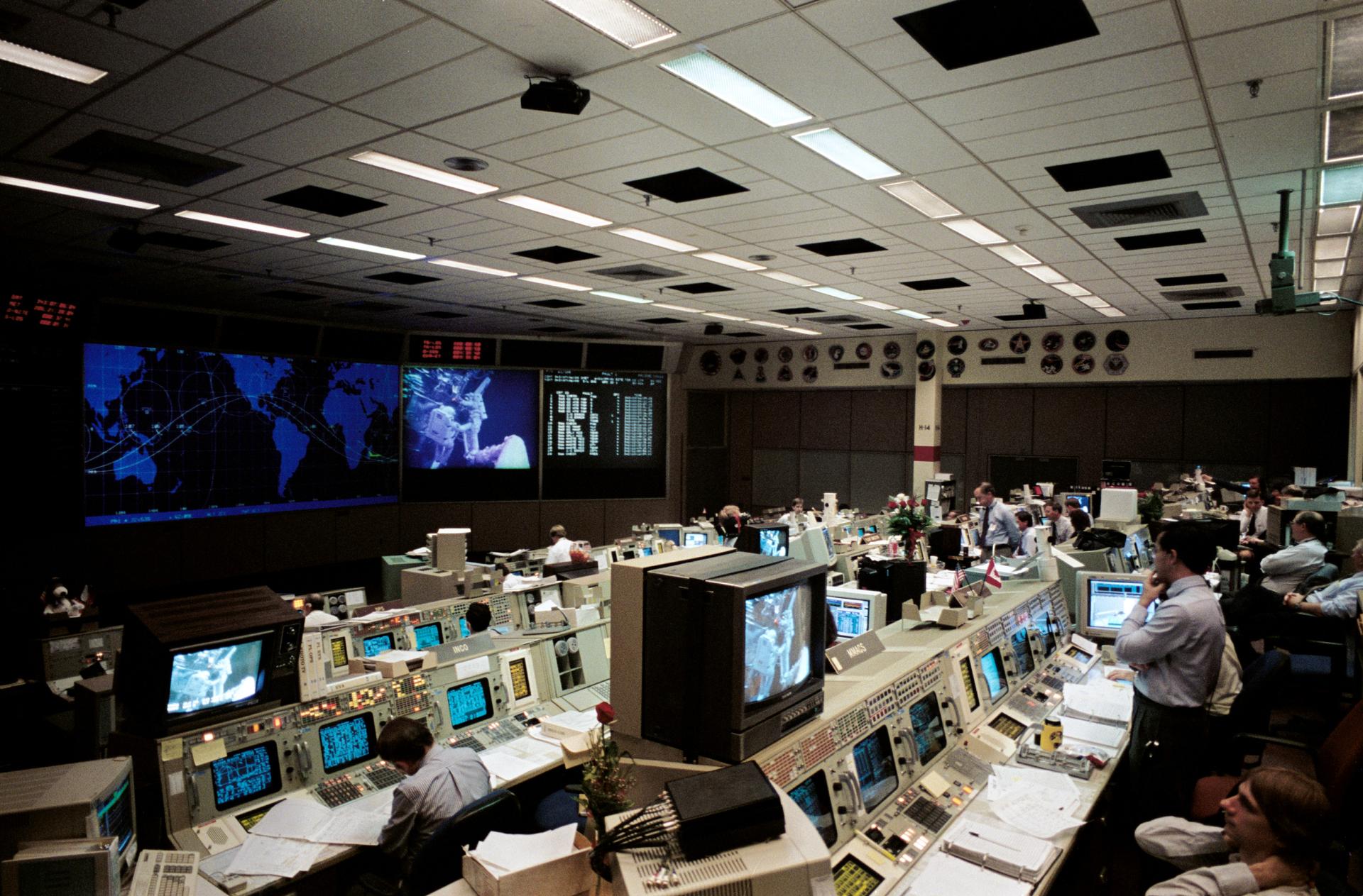 The mission control room at Johnson Space Center is shown with flight controllers watching a Servicing Mission 1 spacewalk.