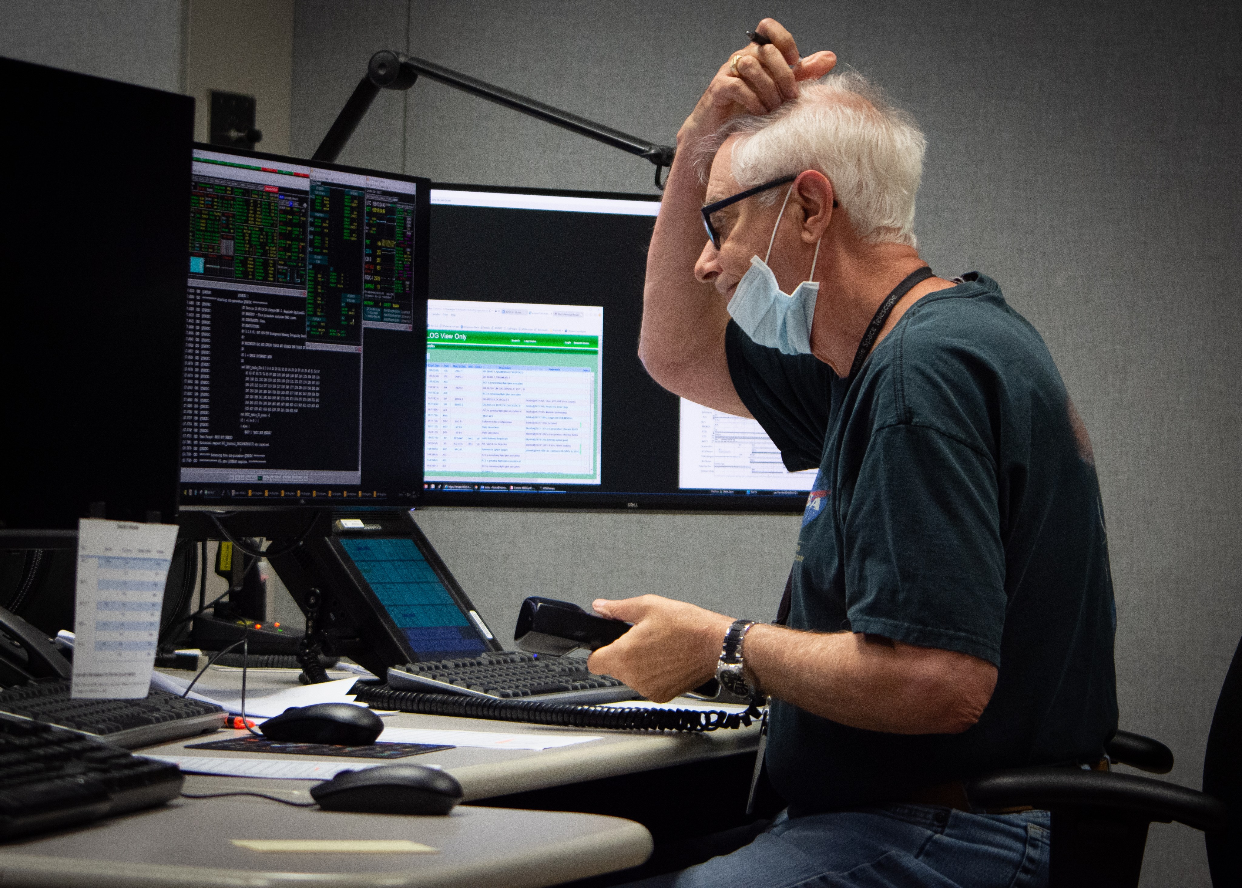 A man scratches his head in thought while holding a land line phone receiver in one hand and analyzing data on a screen in the Hubble control center.