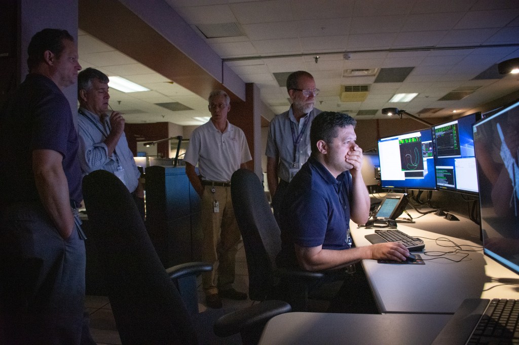 Four people standing discuss the pointing stability of Hubble as another person sits at a console in the Hubble control center.