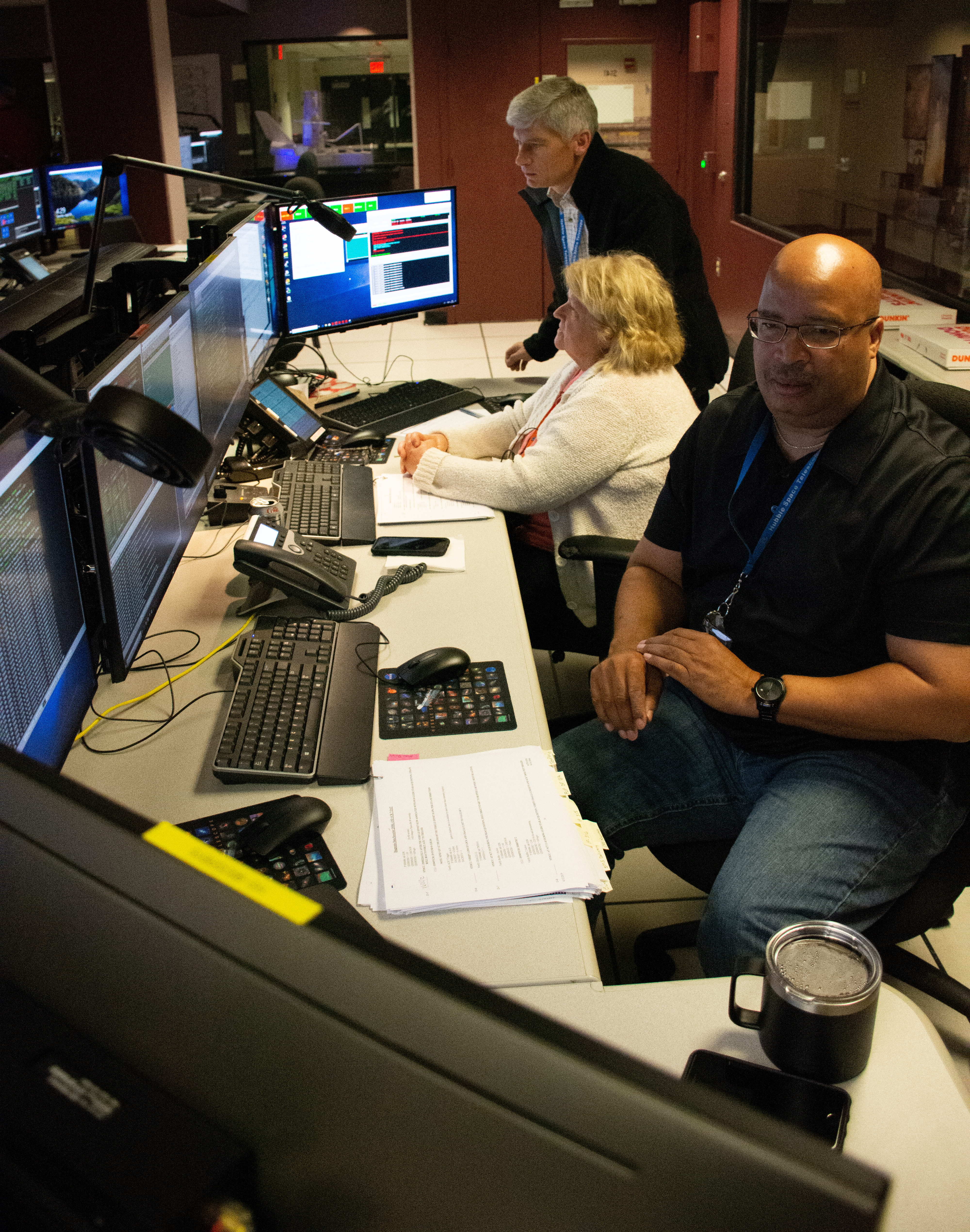 Two people sitting and one standing while monitoring a spacecraft procedure test in the operations area of Hubble.