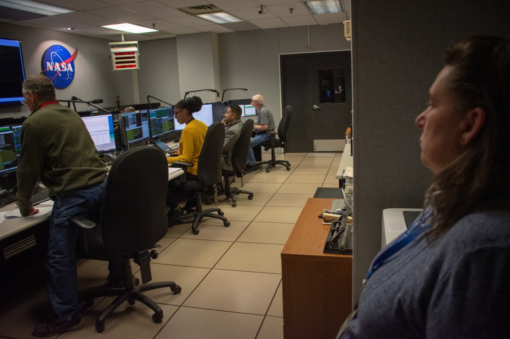 A woman looks on as four flight controllers command Hubble in the control center.