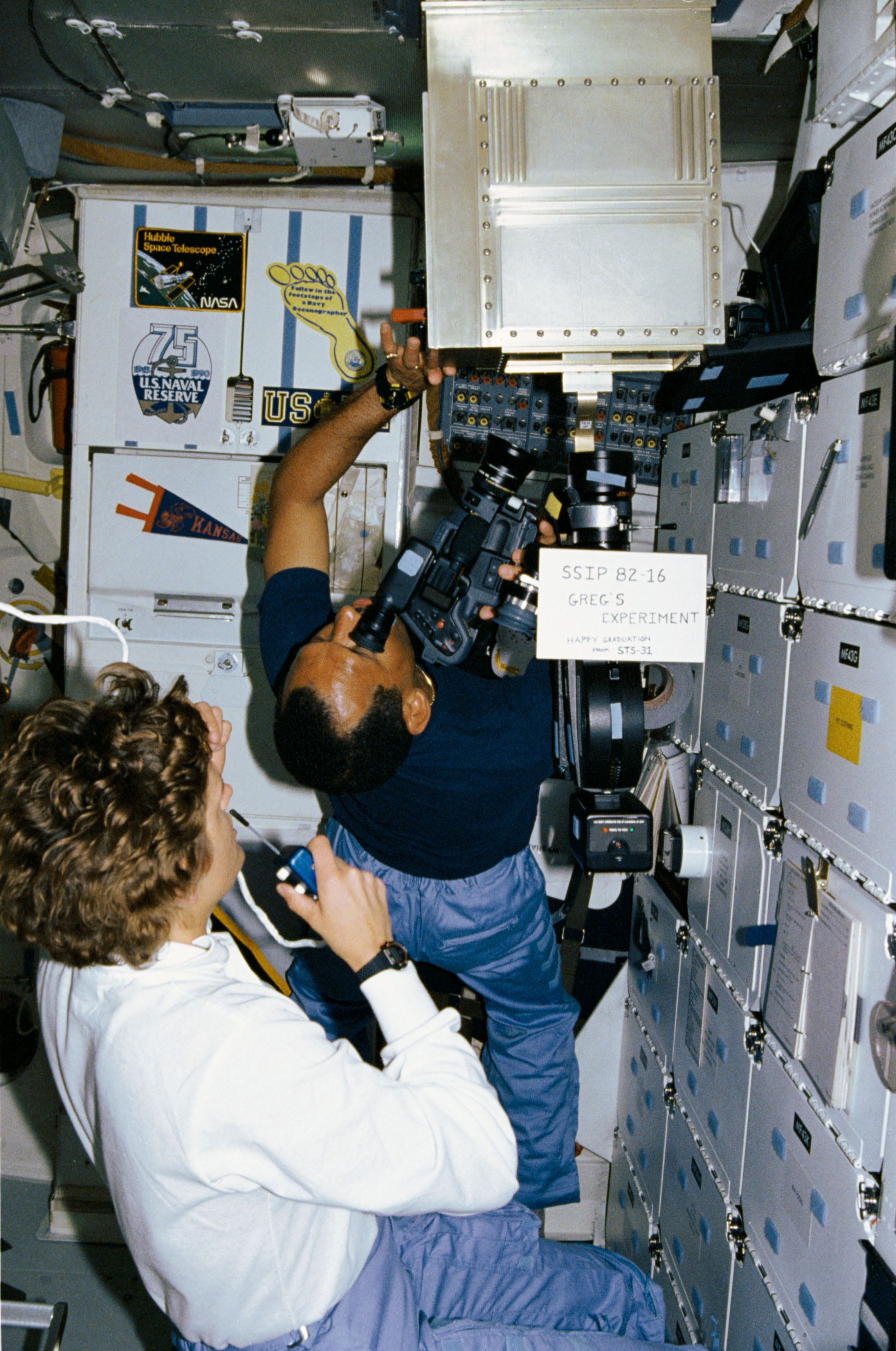 AN astronaut with a video camera and a second astronaut assisting in the space shuttle.