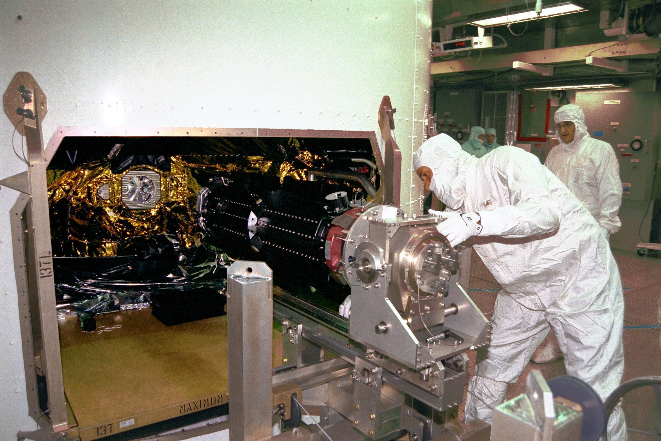 Engineers in cleanroom suits insert a cylindrical device into a spacecraft.