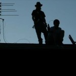 Silhouettes of two men on a rooftop with a radio antenna to their left.