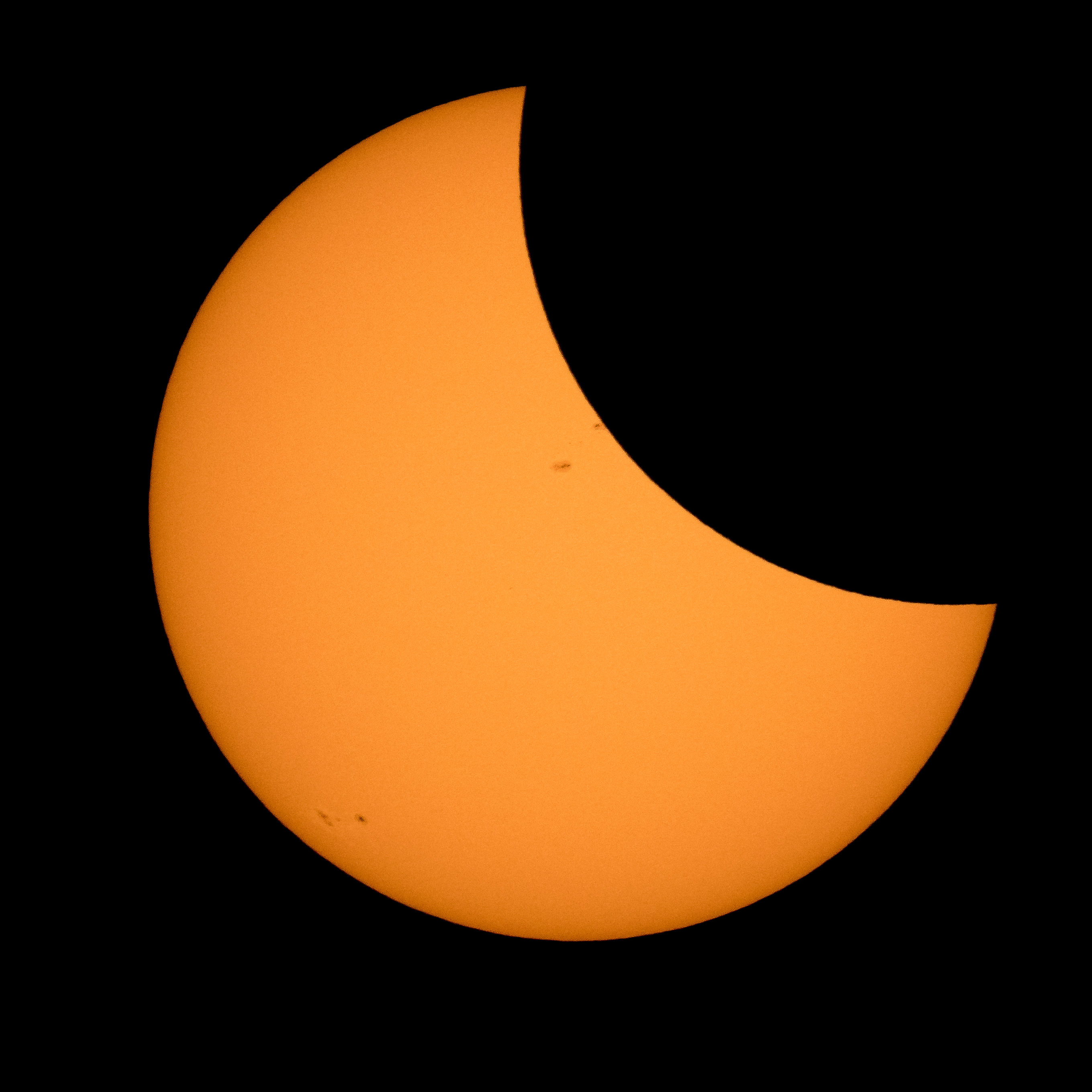 The Sun appears as a large orange crescent, with the upper right portion of it covered by the Moon, which just appears as a black disk. A few sunspots can be seen near the middle and lower left edges of the Sun.