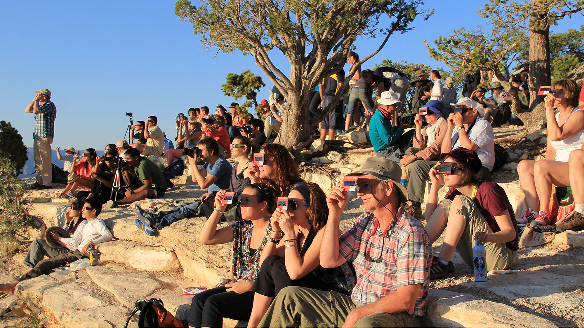 Dozens of people sit or stand outside on a rocky slope and all face the same direction (left) while holding card shaped solar viewers or while wearing solar eclipse glasses. It is a sunny day with a blue sky and trees in the background.