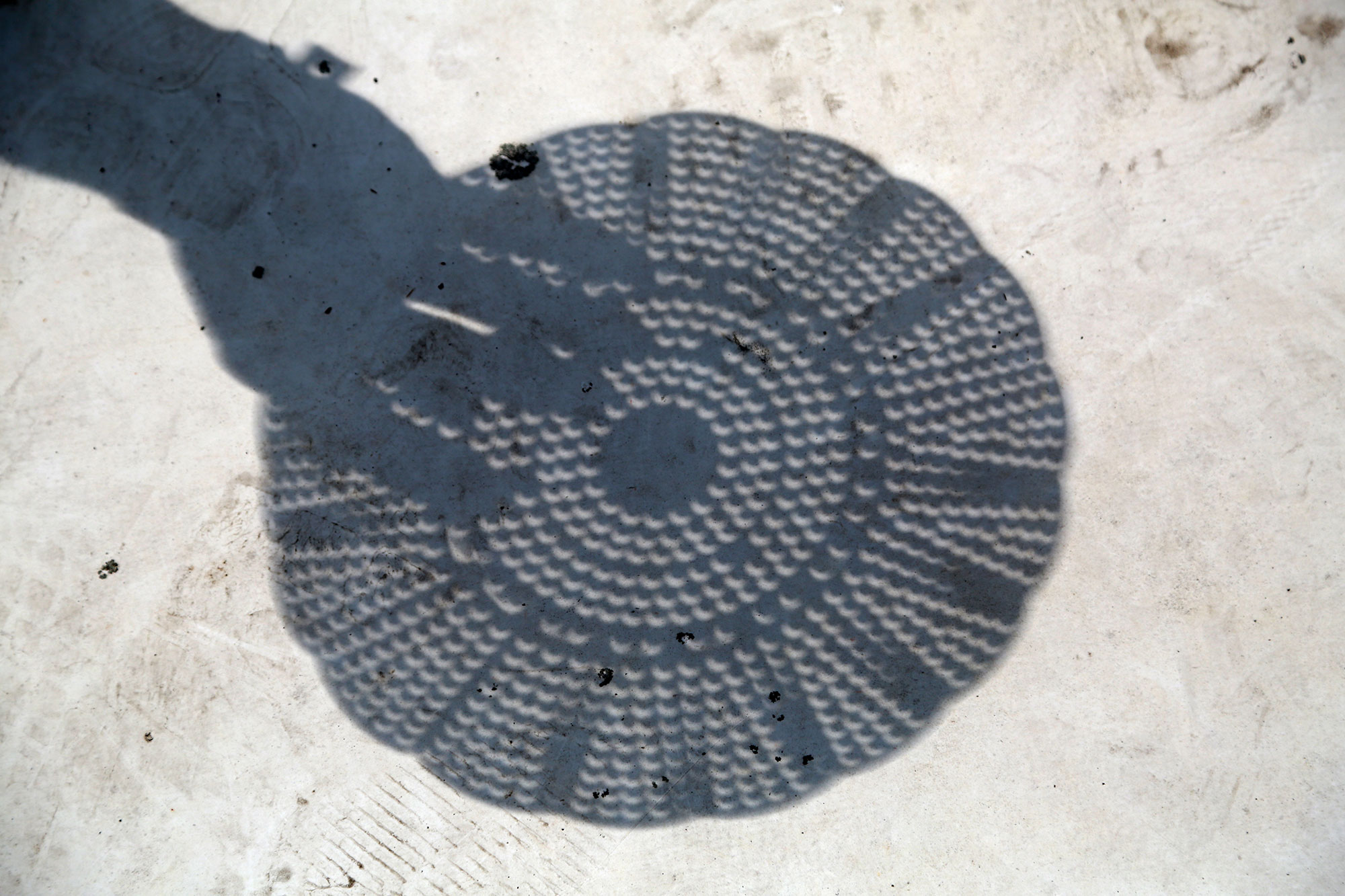 A shadow of a hand and a kitchen colander (which appears as a circle with holes) appears on a white surface. Close inspection reveals that where sunlight passes through the holes of the colander, small crescent shapes appear.