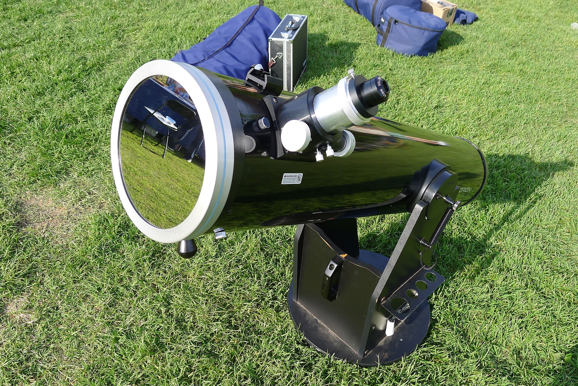 A black telescope sits on grass. On the left end of the telescope, which is pointed slightly upward, is a round cover that reflects a chair and table in the distance.