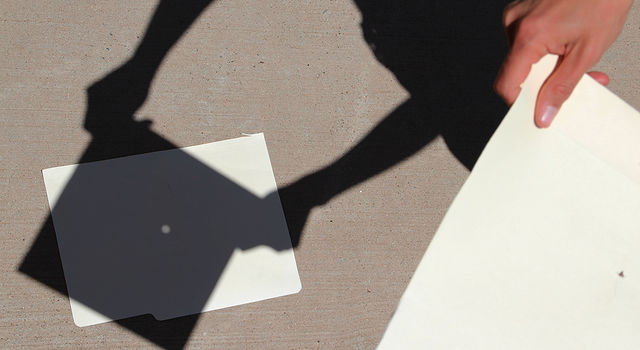 在右边，一张中间戳了一个小洞的白纸占据了图像。一只手拿着纸。在左边，地面上有一个纸的影子。在中间，一个小圆的光穿过——代表太阳。