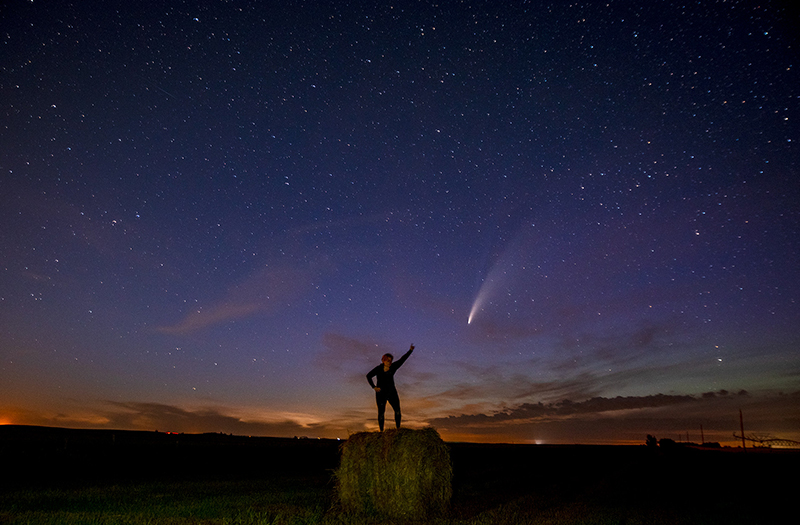 Christy in front of a purple and orange sky, striking a pose and pointing to a comet with a long tail in the sky.