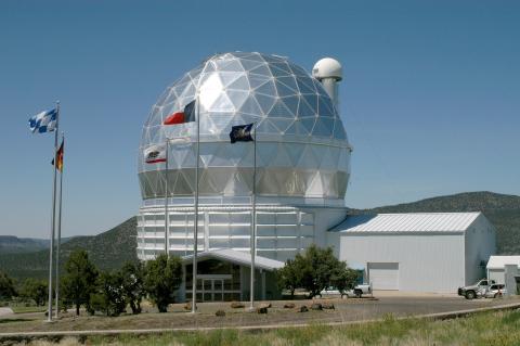 Photo of a large silver domed building with a smaller shed next to it; 3 colorful flags fly in front of the dome.