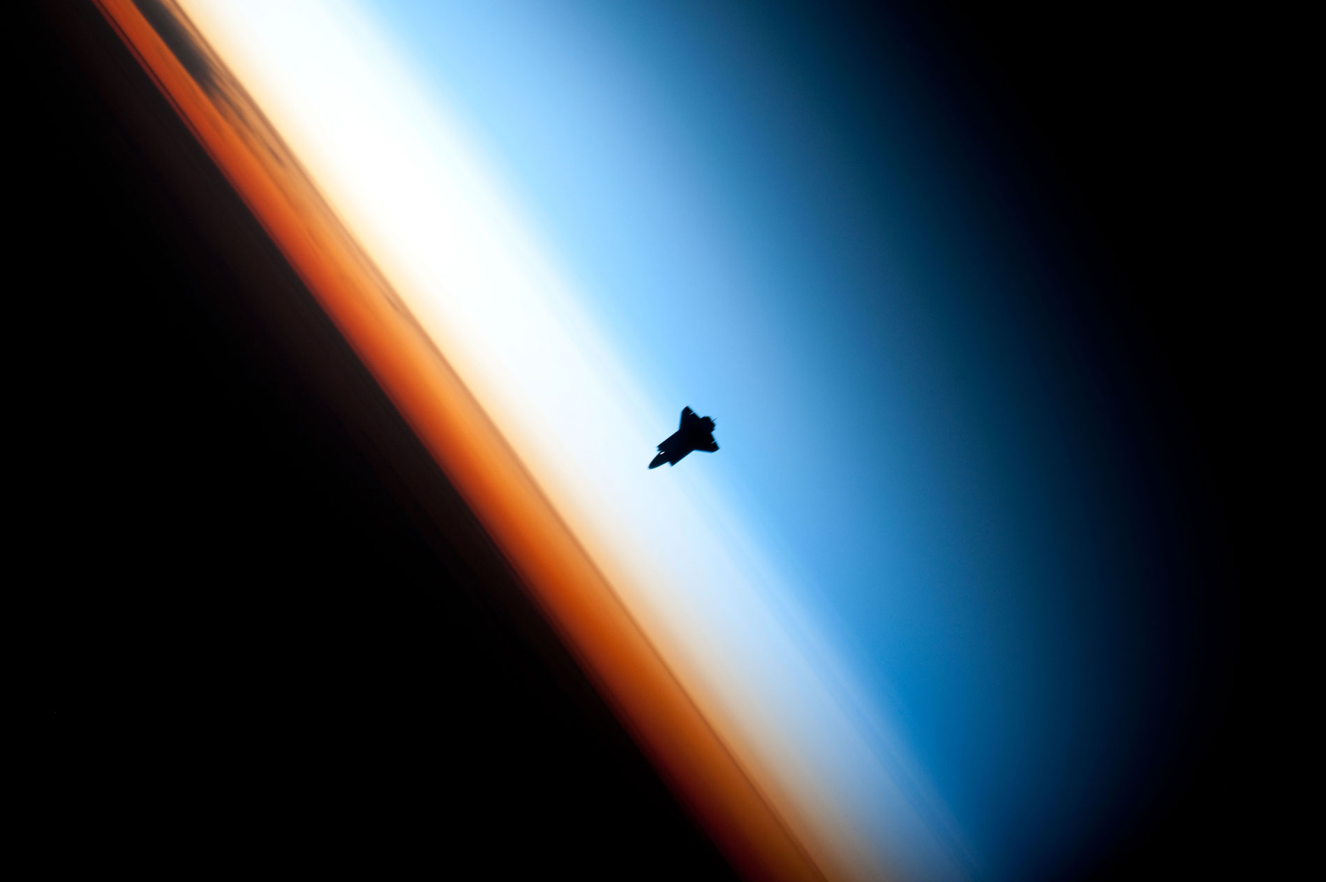 Image of Space Shuttle over a darkened Earth with three colorful layers of atmosphere visible.