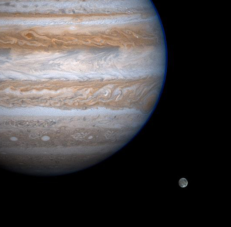 Giant Jupiter with its reddish brown and white swirls looms large over the moon, Ganymede - which looks like a small gray and white marble.