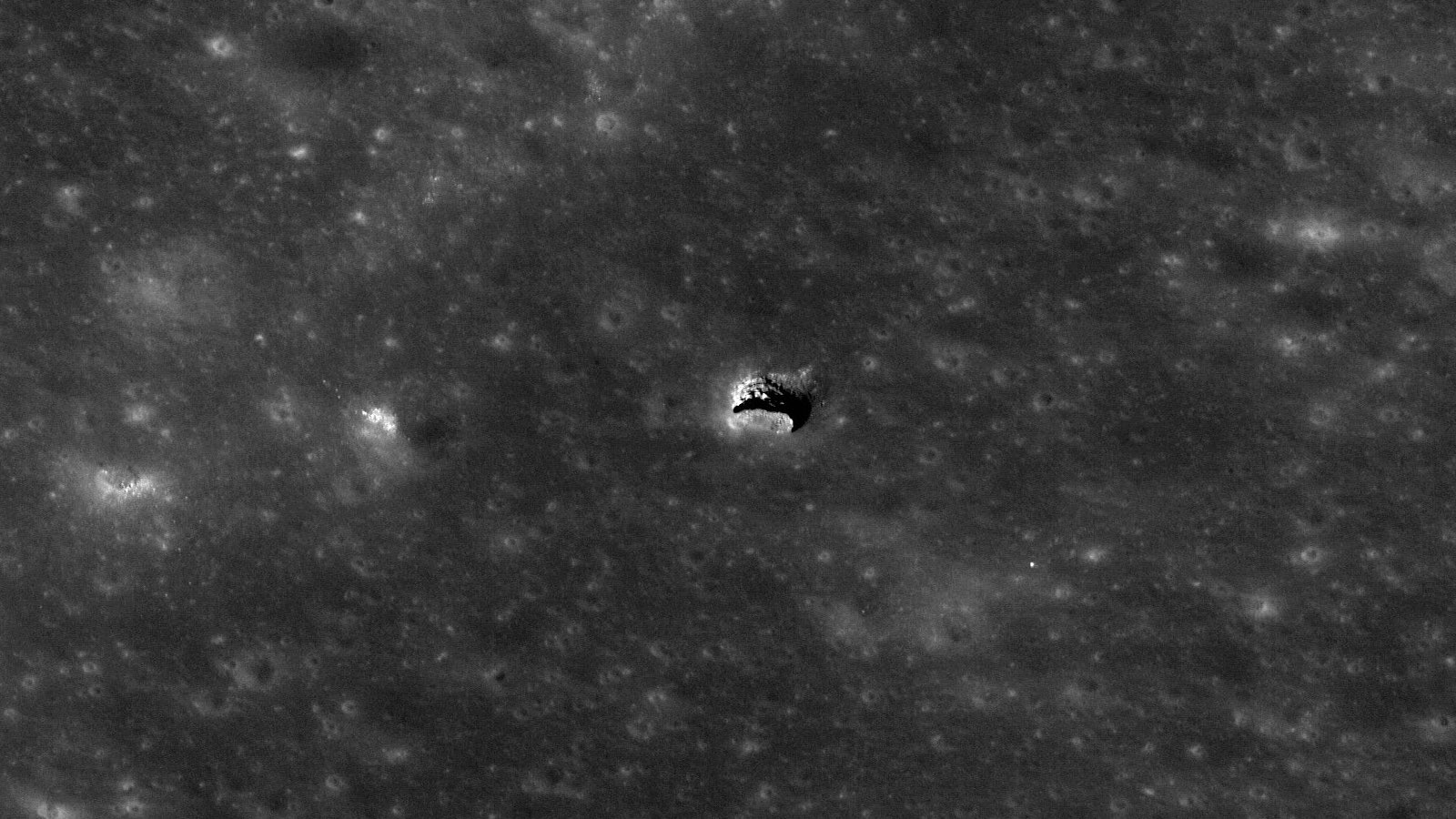 Satellite image of the Moon’s surface. In the middle of the image, the surface is sharply interrupted by what appears to be an opening in the roof of a cave. The landscape is illuminated at a low angle, revealing a small part of what looks like a larger void below the opening.