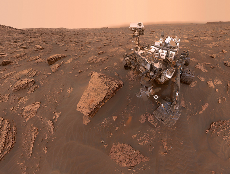 This selfie was taken by NASA's Curiosity Mars rover. Varying shades of red and brown rocks are visible below a dusty atmosphere.