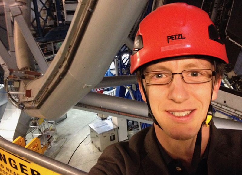 Portrait photo of a smiling man wearing glasses and a red hard hat and standing in front of very large equipment in a lab