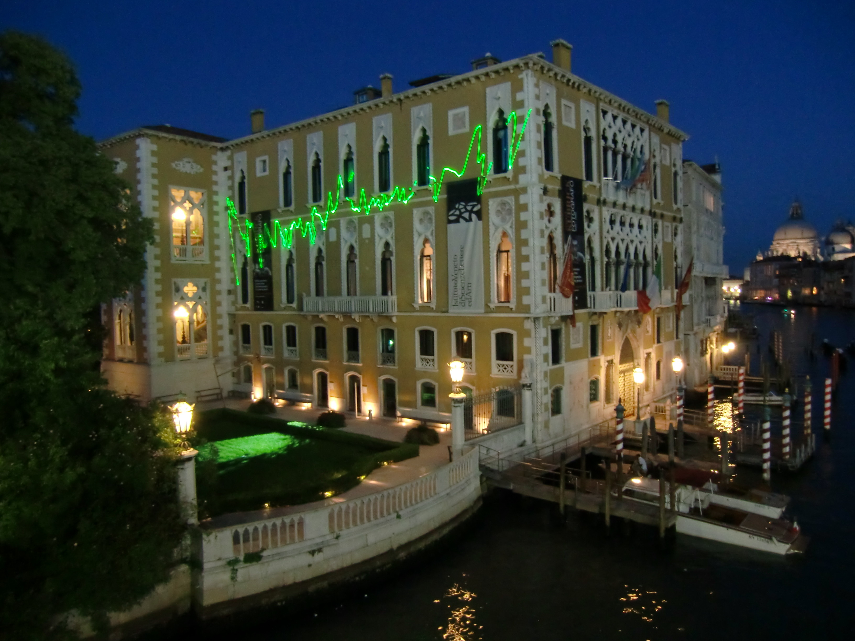 A jagged green line of laser light, resembling an EKG, plays across the face of a yellow and white building on the Venice canals at night.