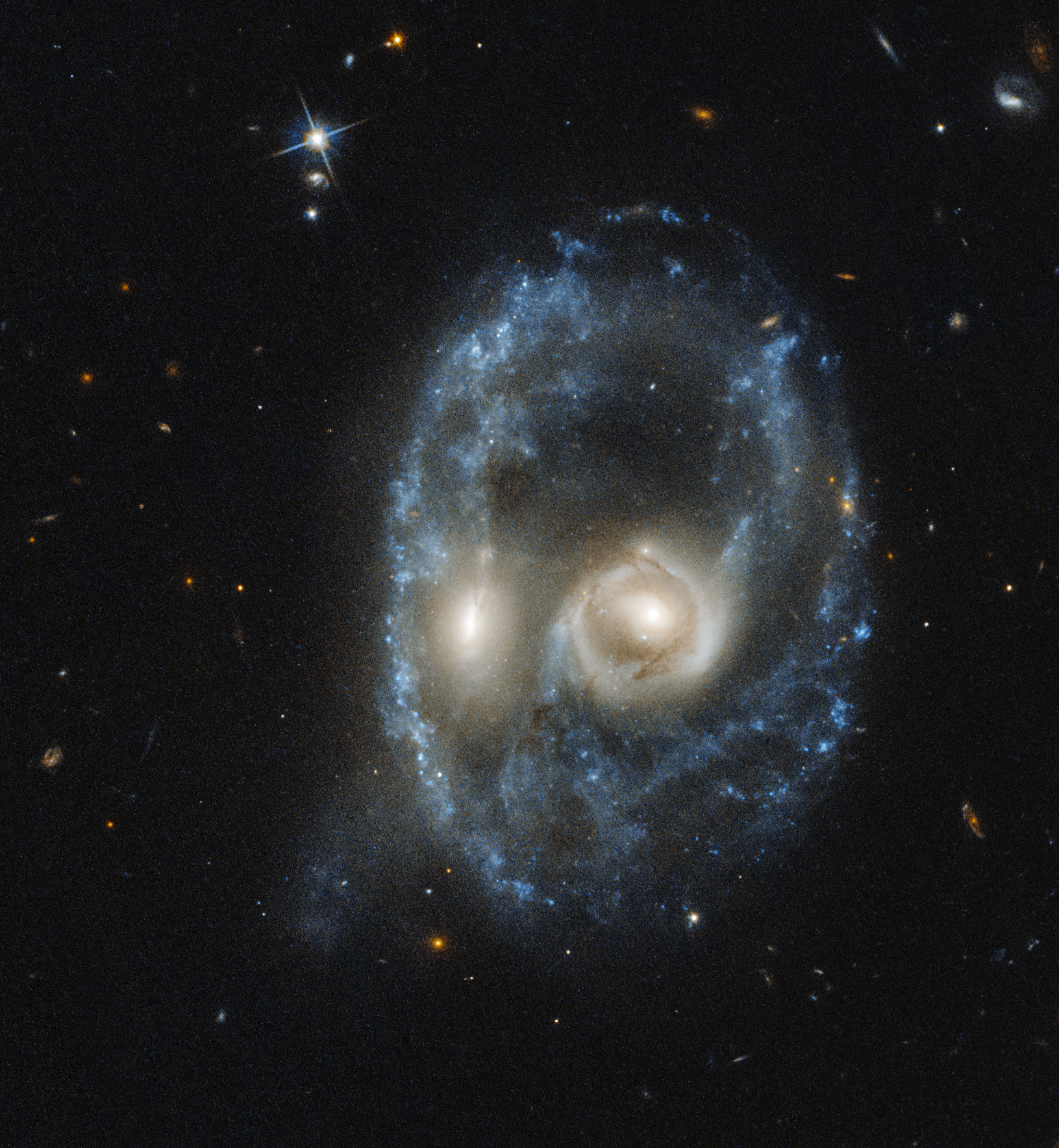Two galaxies, each has a bright-white core. Their cores are very close together. They are surrounded by light blue gas, dust, and stars that form a ring around the cores.