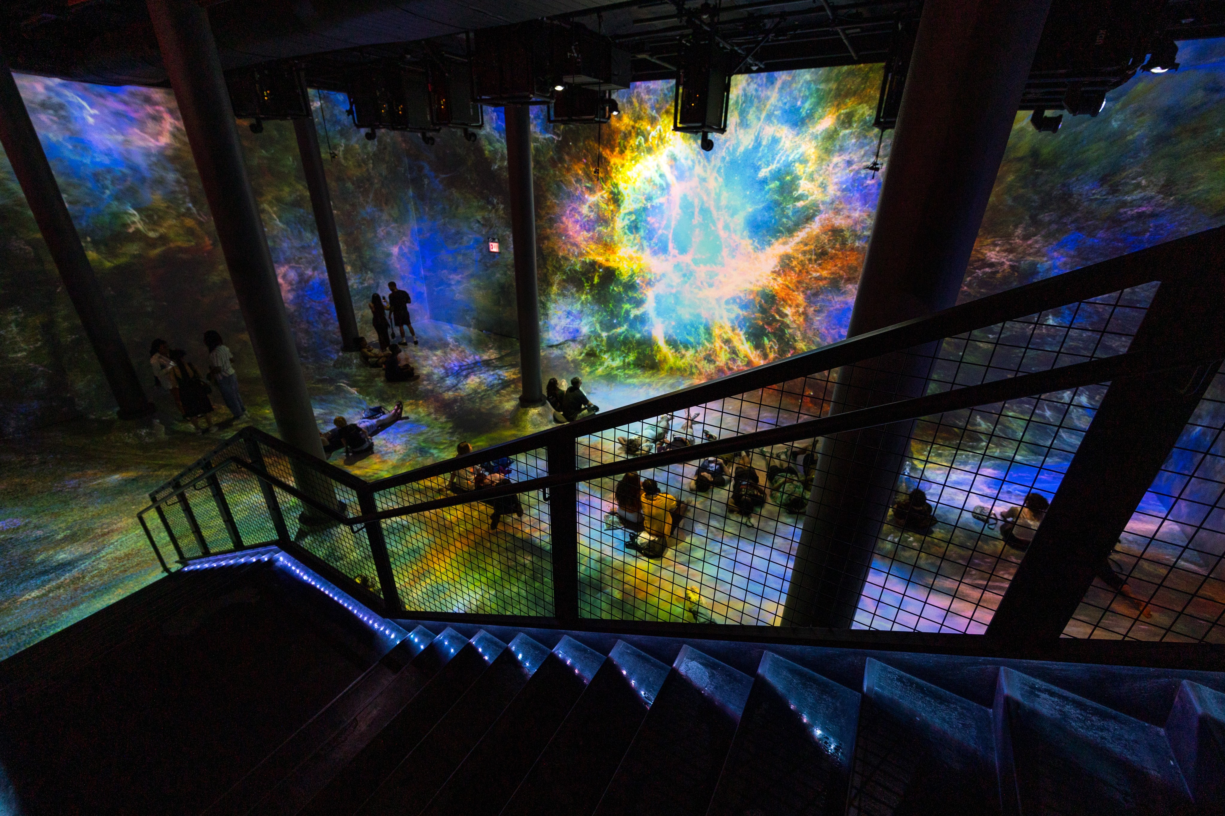 People sit and stand at an art exhibit in a dark room surrounded by extremely tall projected Hubble images.