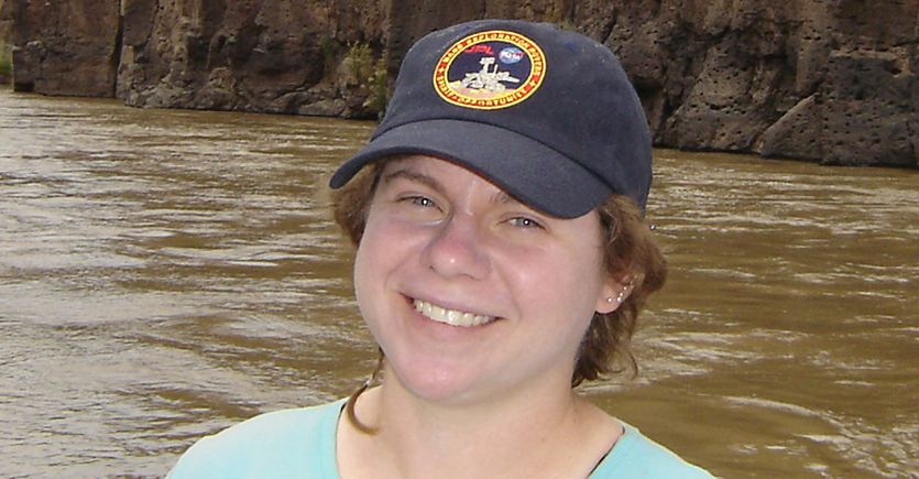 A portrait photo of NASA scientist Barbara Cohen. She's standing in front of a body of water and wearing an hat.