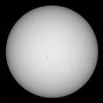 The Sun, shown as a bare orb in black and white. Some sunspots speckle the gray surface in black.